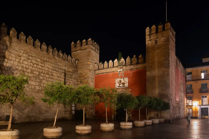 Puerta del León del Real Alcázar de Sevilla de noche, con la muralla almenada iluminada y el azulejo del león sobre la entrada