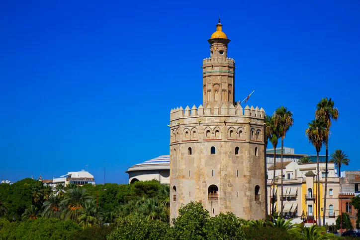 La Torre del Oro de Sevilla con sus tres cuerpos, almenas y cúpula dorada, entre palmeras bajo un cielo azul intenso