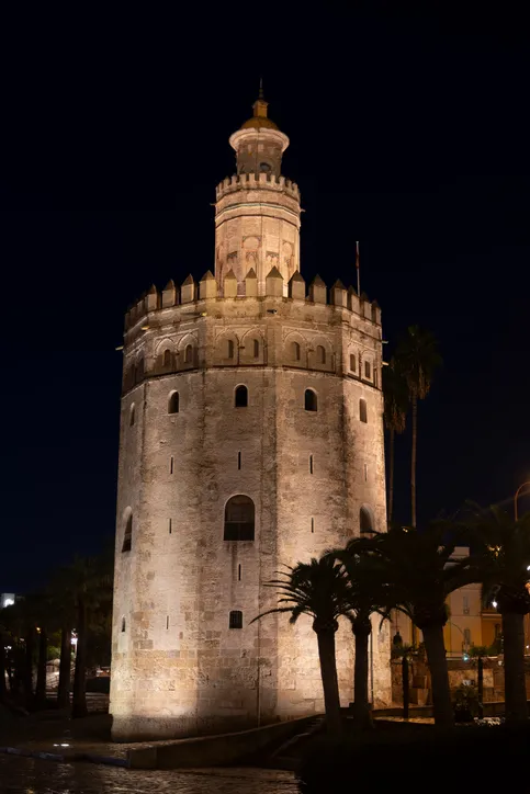 La Torre del Oro iluminada de noche, con sus almenas y palmeras a los pies, sede del Museo Naval de Sevilla