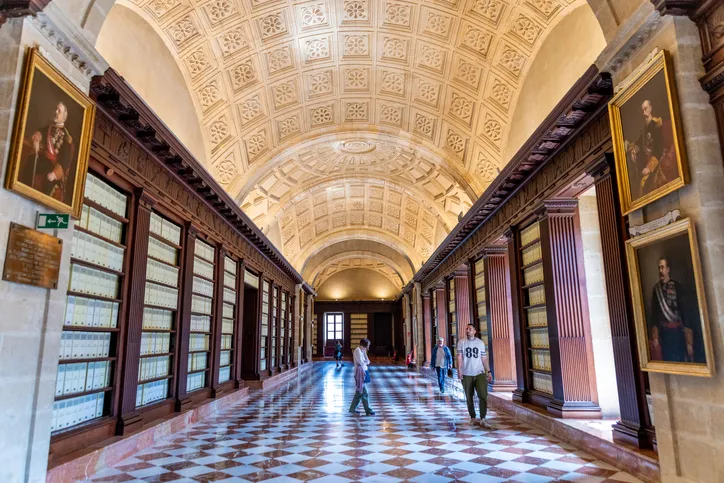 Galería del Archivo de Indias con estanterías de madera, retratos, techo abovedado y suelo de mármol ajedrezado