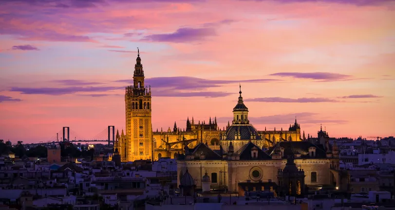 La Catedral de Sevilla y la Giralda iluminadas al atardecer bajo un cielo rosado, sobre los tejados del centro histórico